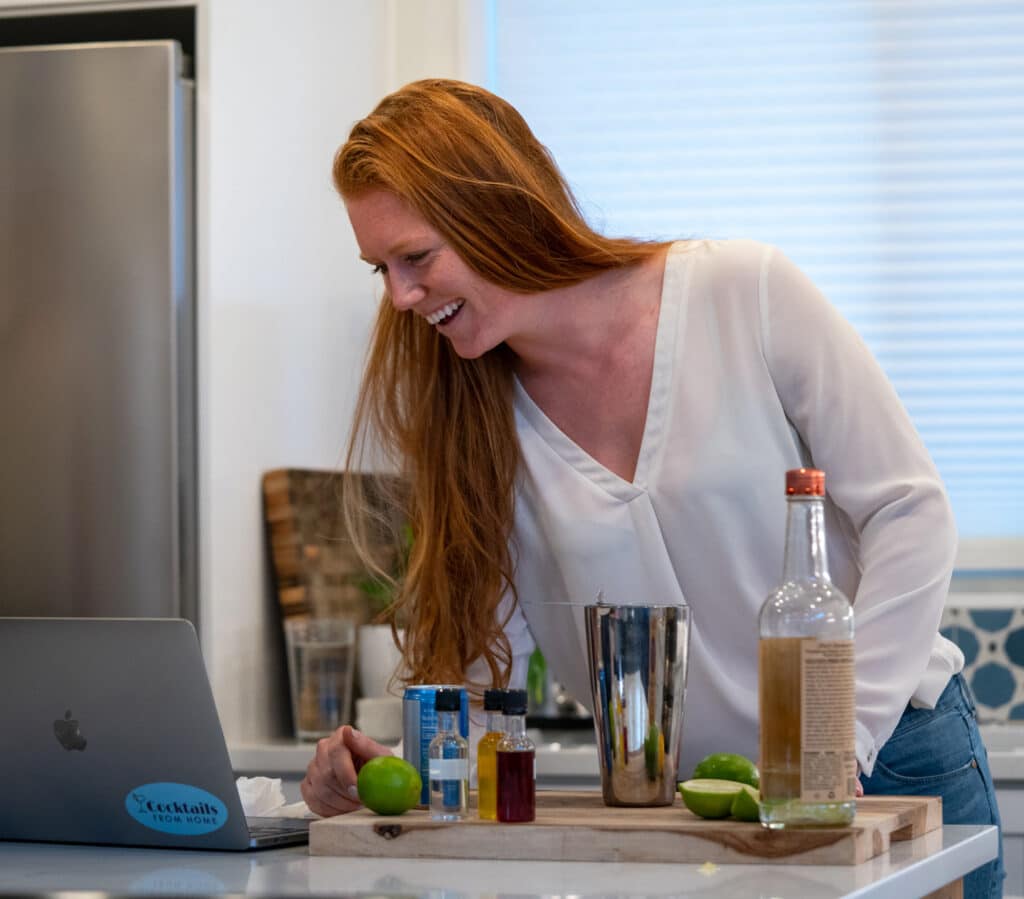 A smiling woman stands at a counter with bartending supplies looking at an open laptop during a virtual happy hour.