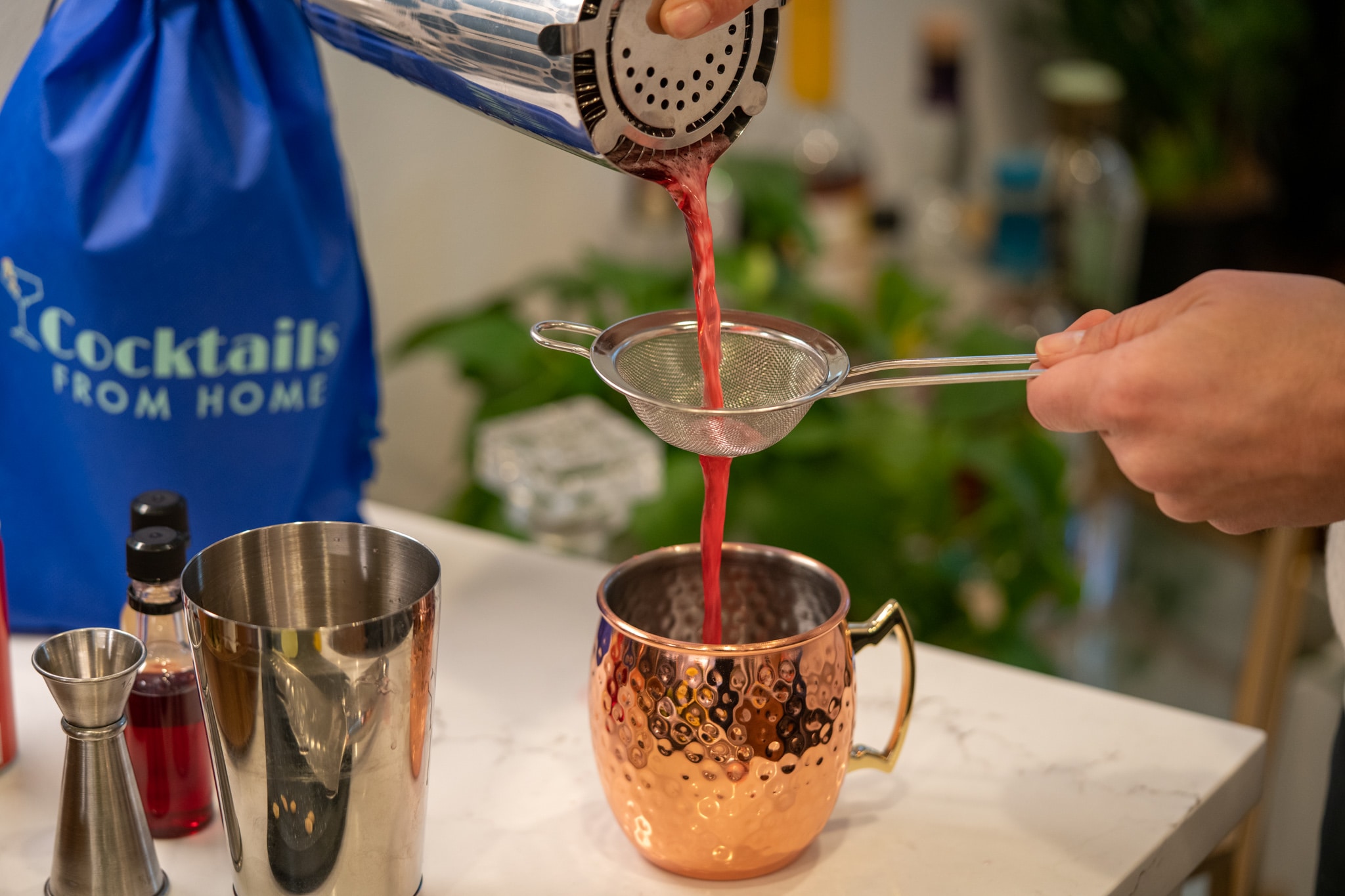 The contents of a cocktail shaker are poured through a mesh strainer into a copper Moscow Mule mug.